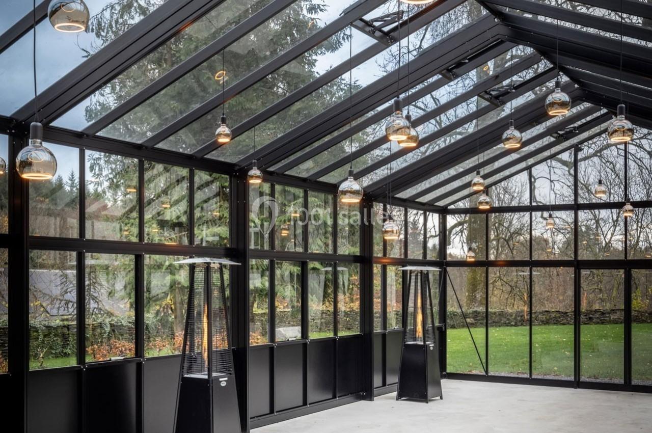 Intérieur d'une véranda moderne en verre et métal, avec des lampes suspendues et vue sur un jardin arboré.