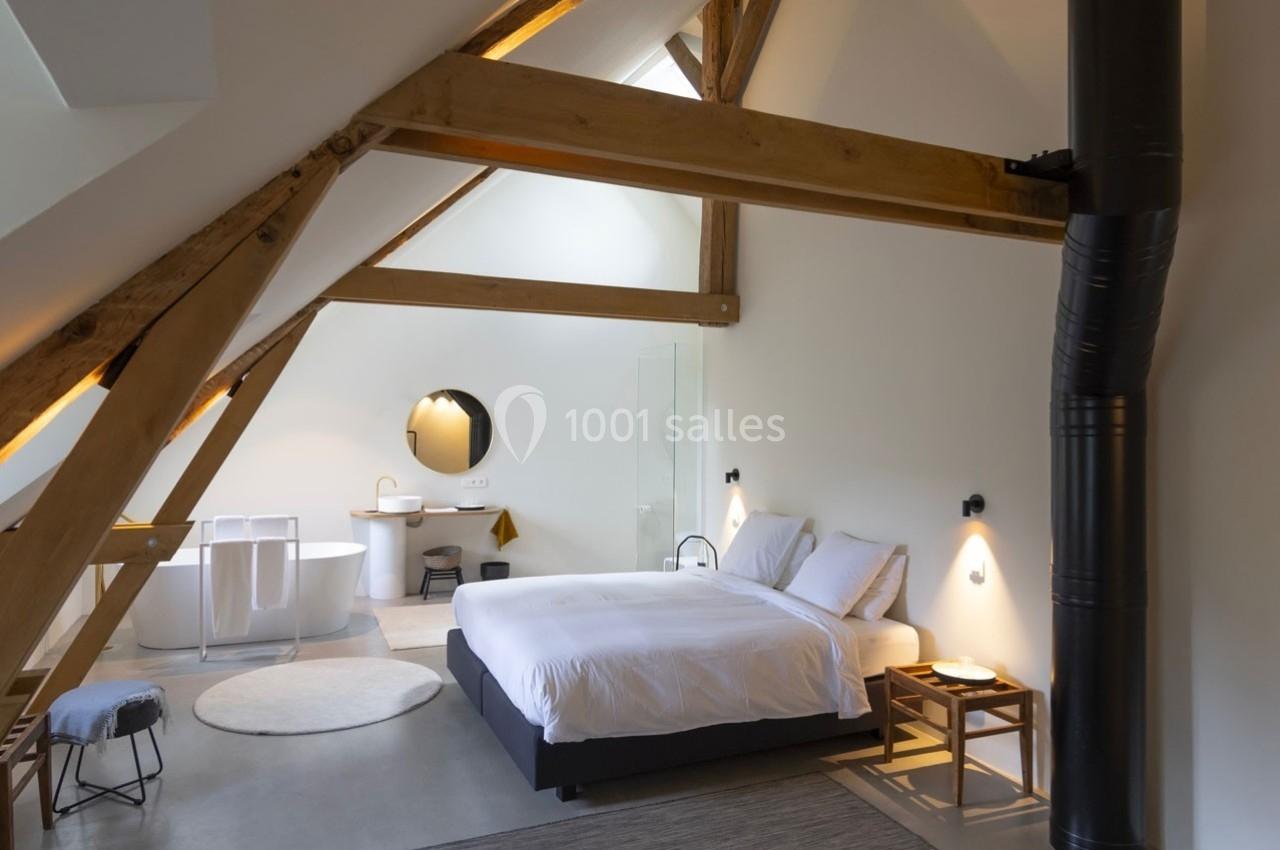 Chambre mansardée lumineuse avec poutres apparentes, lit double, baignoire, lavabo et poêle noir.