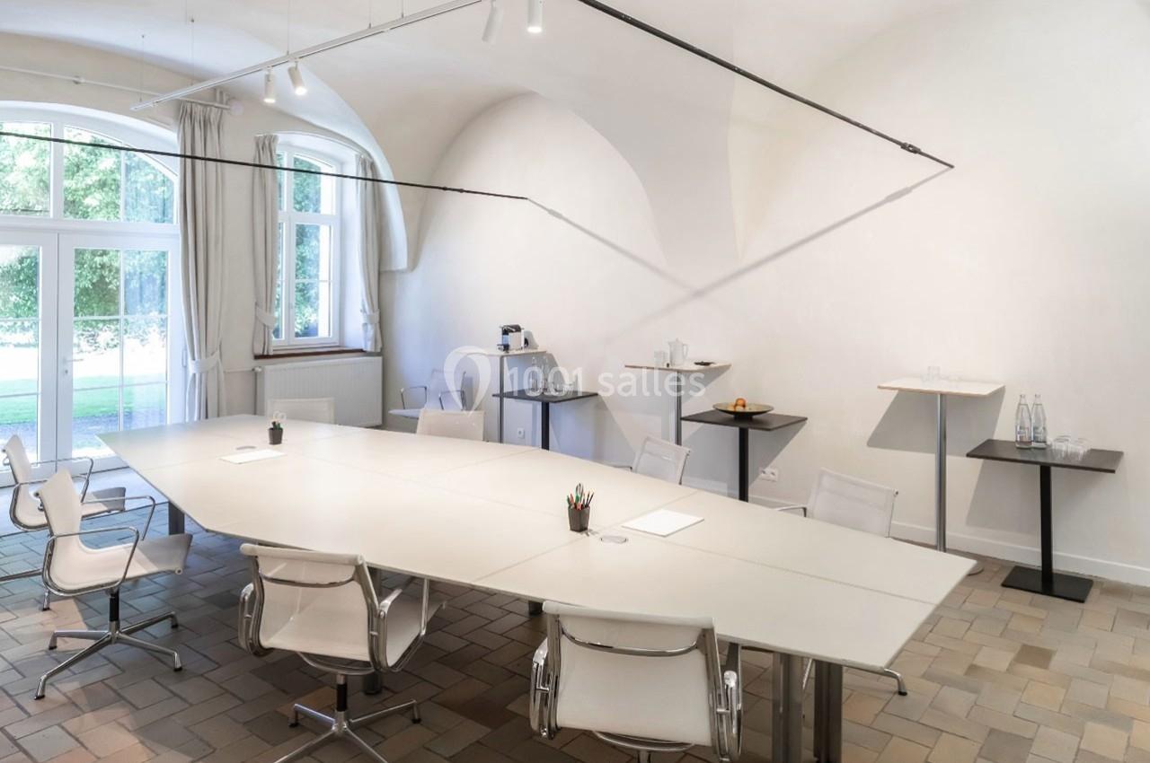 Salle de réunion lumineuse avec une grande table blanche, des chaises modernes et des fenêtres donnant sur un espace vert.