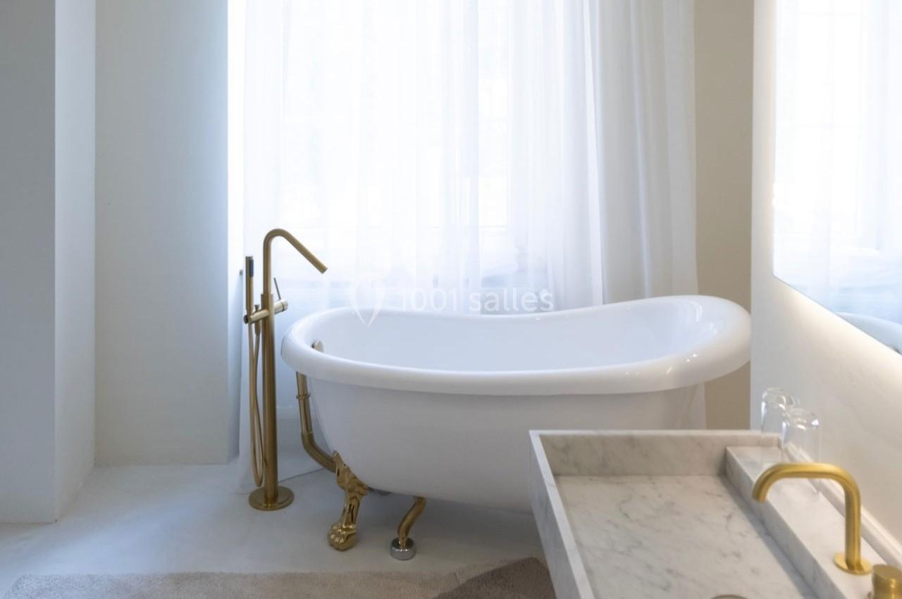 Salle de bain lumineuse avec baignoire sur pieds, robinetterie dorée et lavabo en marbre devant une grande fenêtre.