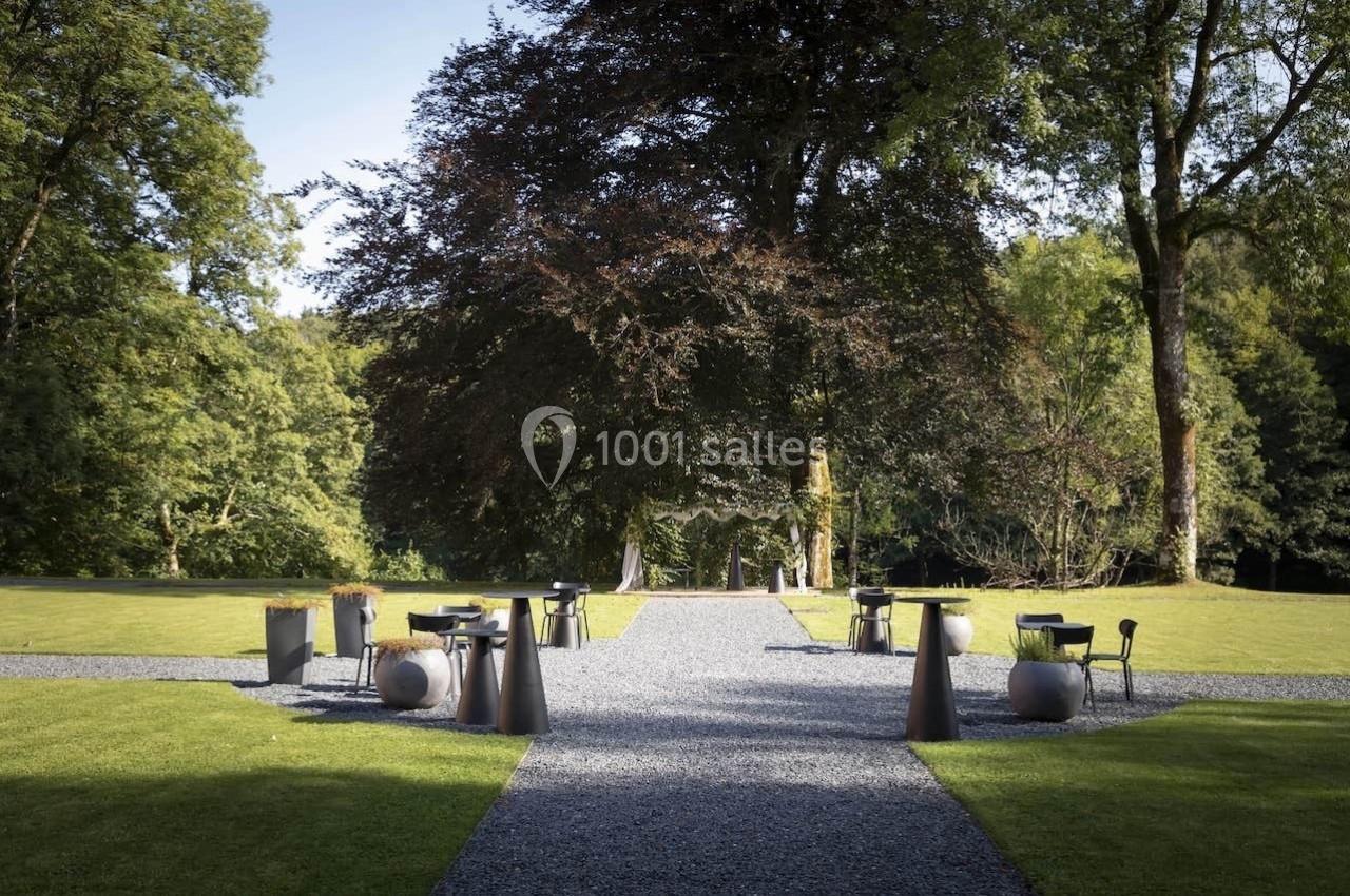 Allée en gravier bordée de pelouses, avec tables et chaises disposées sous de grands arbres dans un cadre naturel.