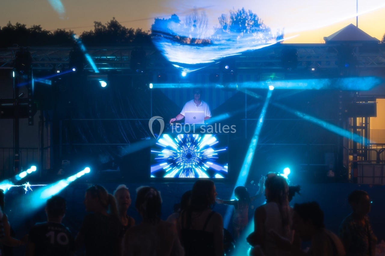 Scène de concert en plein air avec un DJ sur scène, des lumières colorées et une foule dansant au crépuscule.
