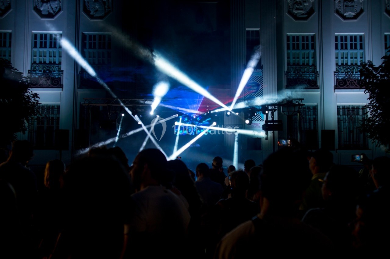 Foule devant un bâtiment illuminé, avec des faisceaux lumineux colorés traversant la scène lors d'un événement nocturne.