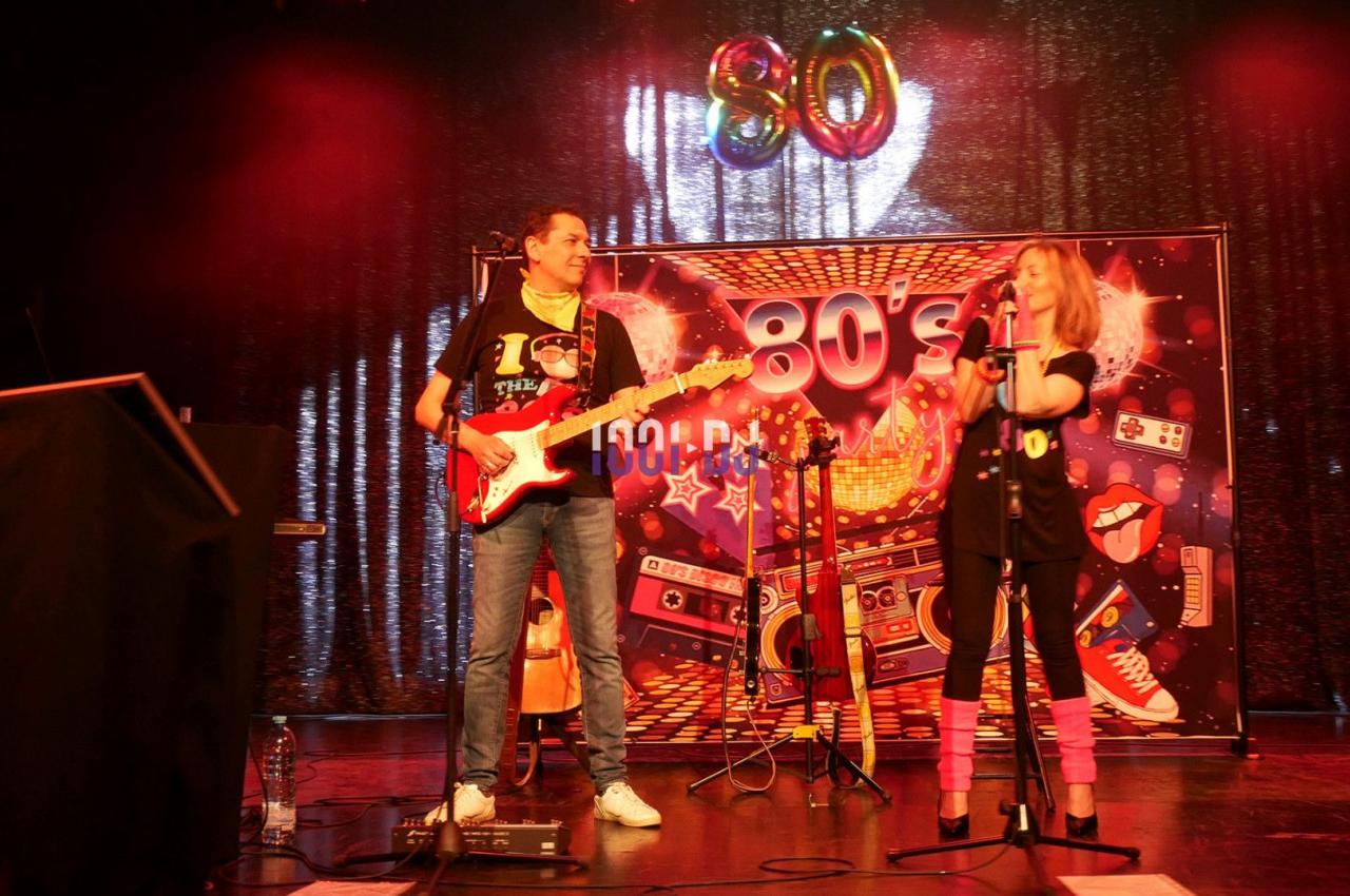 Deux musiciens sur scène jouent de la guitare et chantent devant une décoration sur le thème des années 80.
