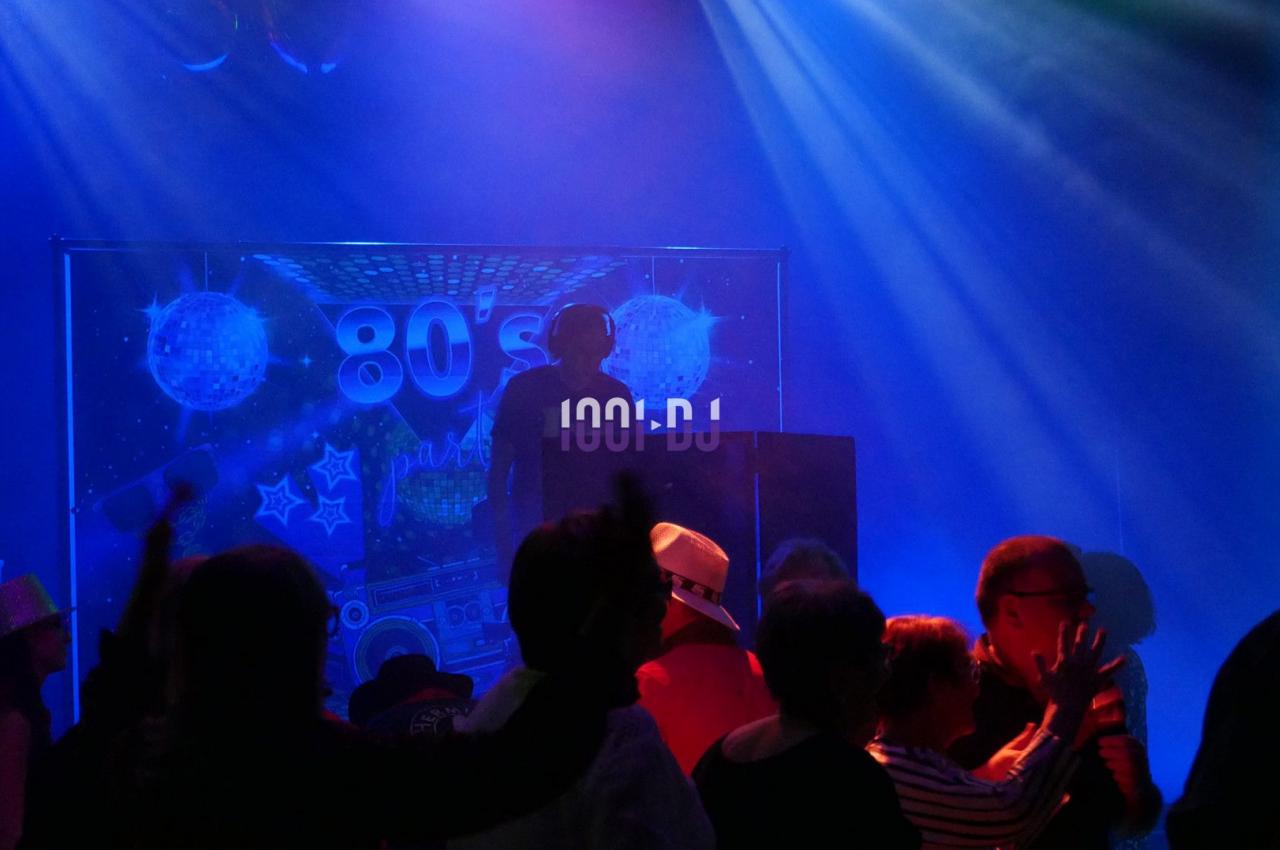 Personnes dansant dans une ambiance de fête rétro avec un DJ devant un décor sur le thème des années 80.