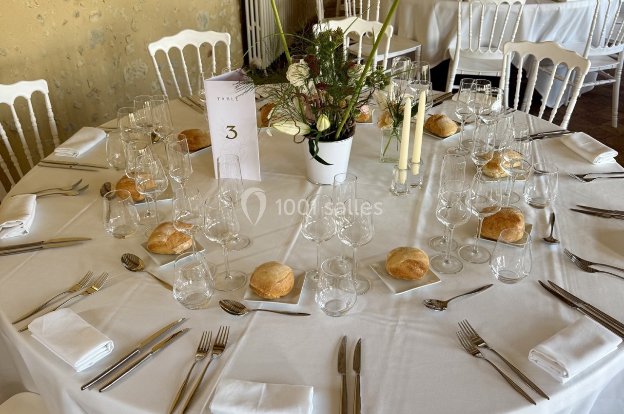 Table ronde dressée avec nappes blanches, couverts, verres, bougies, pain et un centre de table floral.