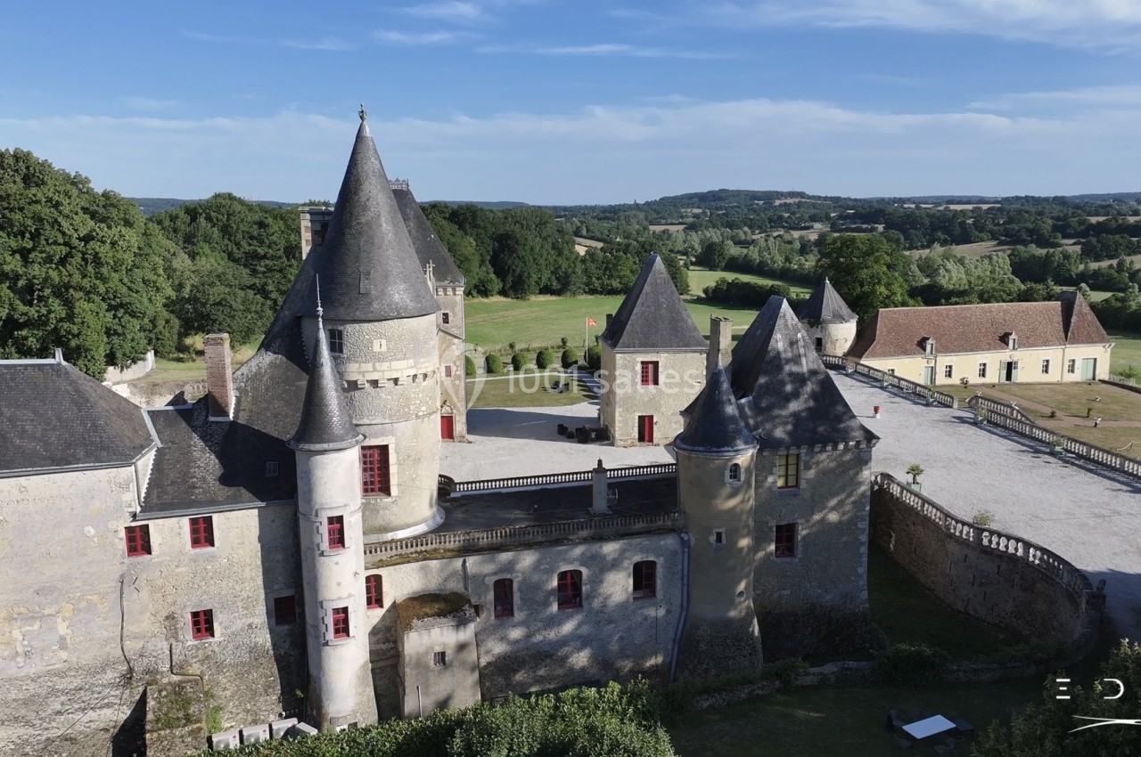Vue aérienne d'un château médiéval avec tours et toits en ardoise, entouré de verdure et d'un paysage vallonné.