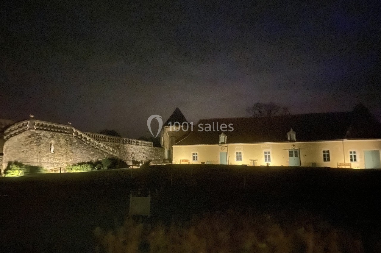 Mur en pierre et bâtiment éclairés la nuit, avec des fenêtres et des portes colorées, dans un cadre sombre.