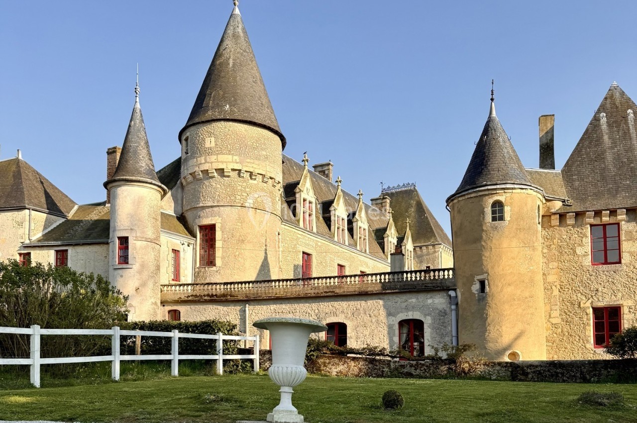 Château en pierre avec plusieurs tours et fenêtres rouges, entouré de verdure et d'une clôture blanche.