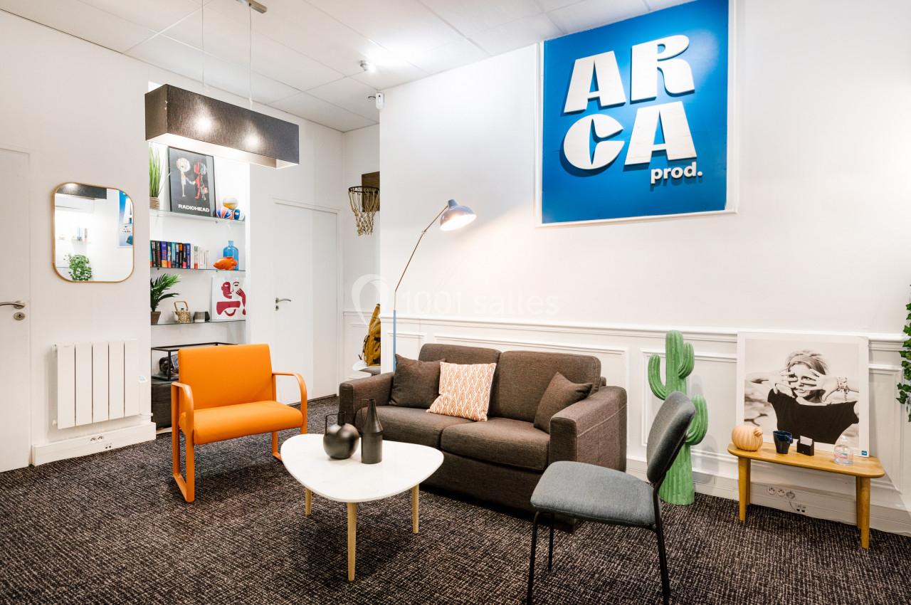Salon moderne avec canapé gris, fauteuil orange, table basse blanche, étagères décorées et logo ’ARCA prod.’ au mur.