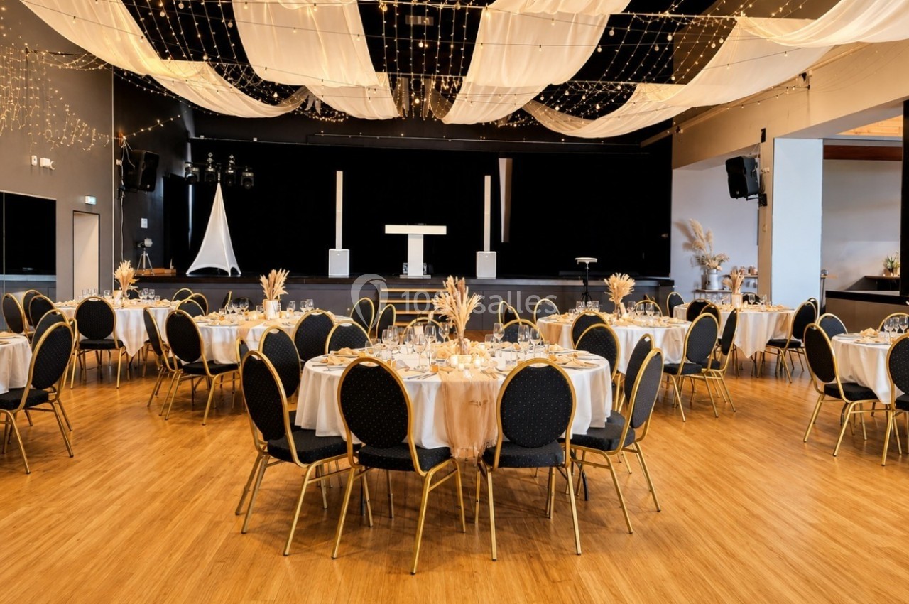 Salle de réception décorée avec des tables rondes dressées, des chaises noires et un plafond orné de guirlandes lumineuses.