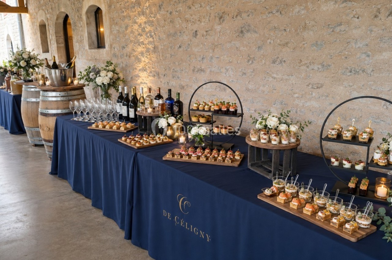 Buffet élégant avec verrines, amuse-bouches, bouteilles de vin et décorations florales sur des tables drapées de bleu.