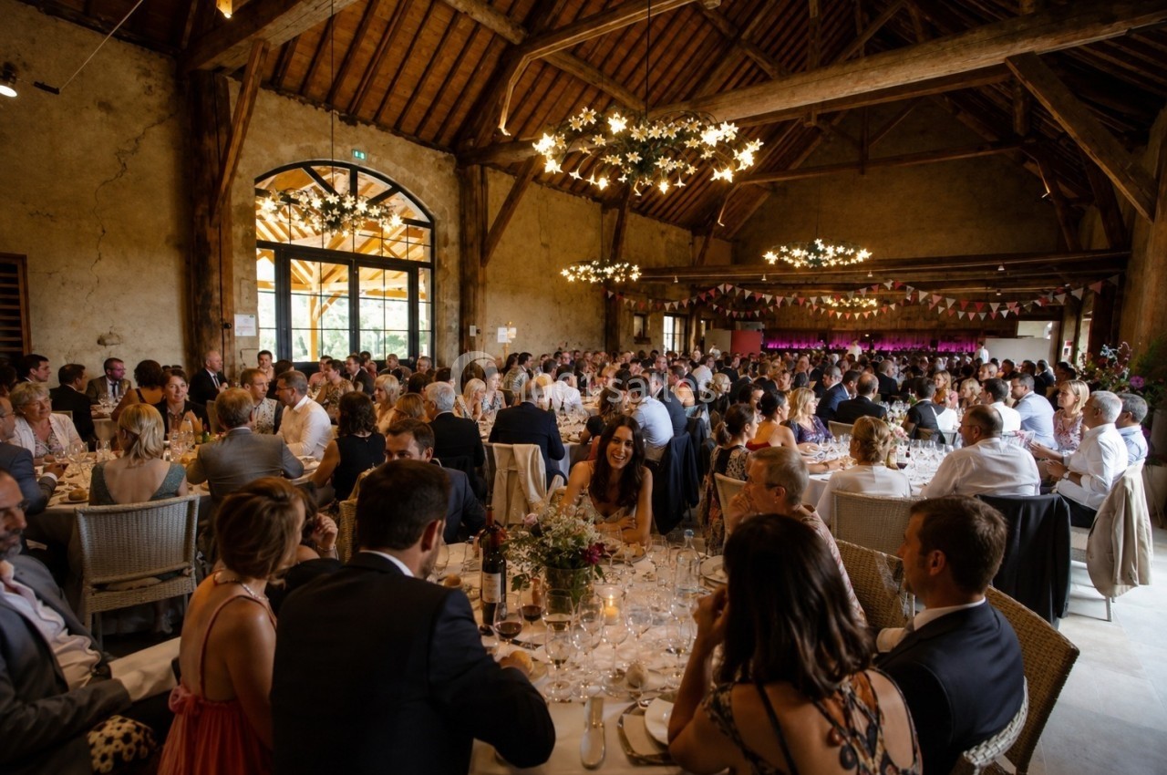Salle de réception rustique avec de nombreux convives assis à des tables rondes, éclairée par des lustres suspendus.