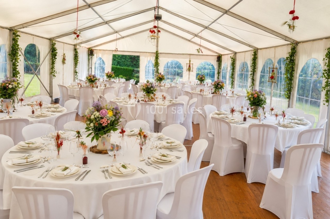 Salle de réception sous une tente avec tables rondes dressées, décorations florales et vue sur un jardin verdoyant.