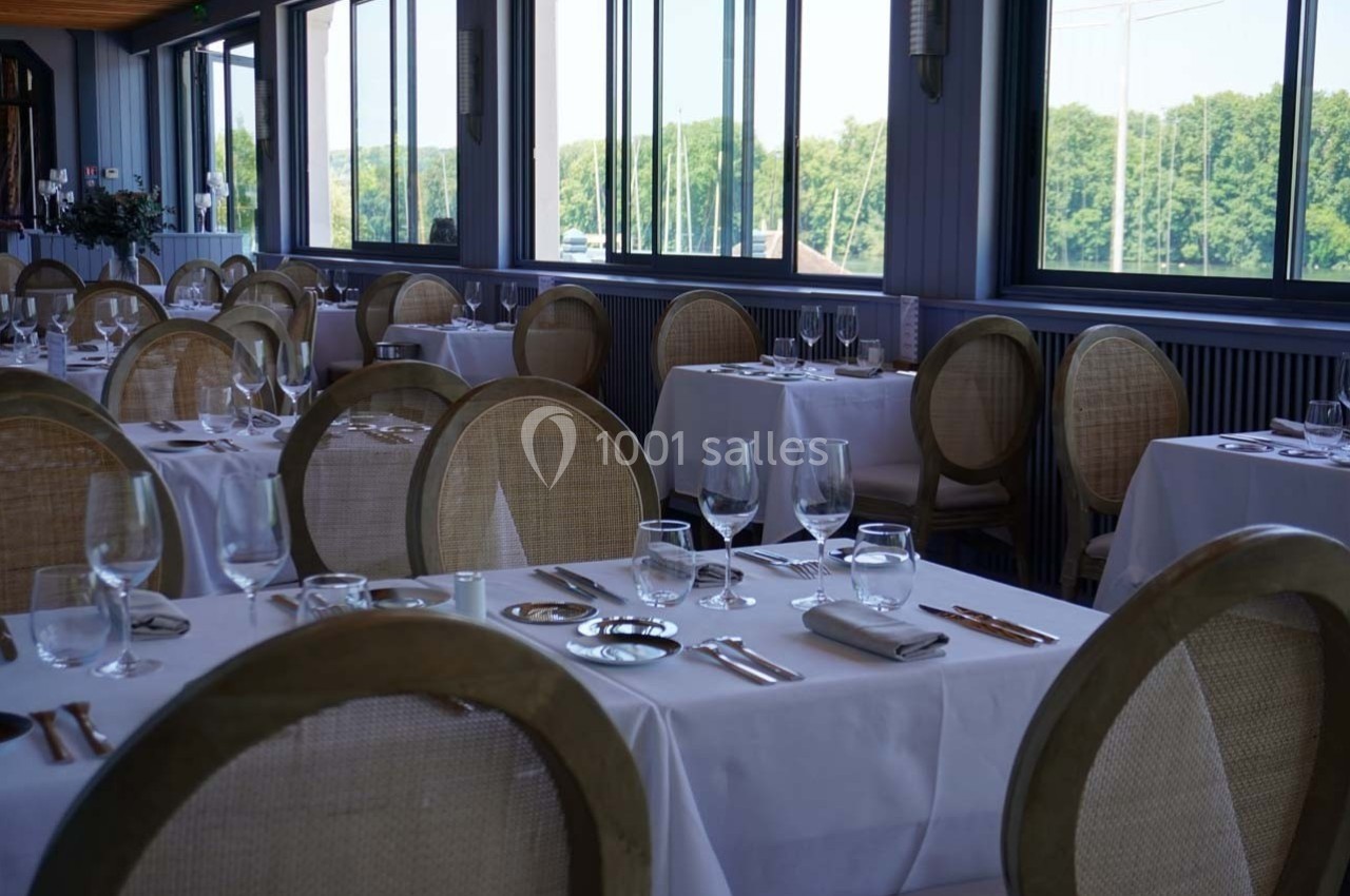 Salle de restaurant élégante avec tables dressées, nappes blanches, chaises en bois et grandes fenêtres donnant sur un…