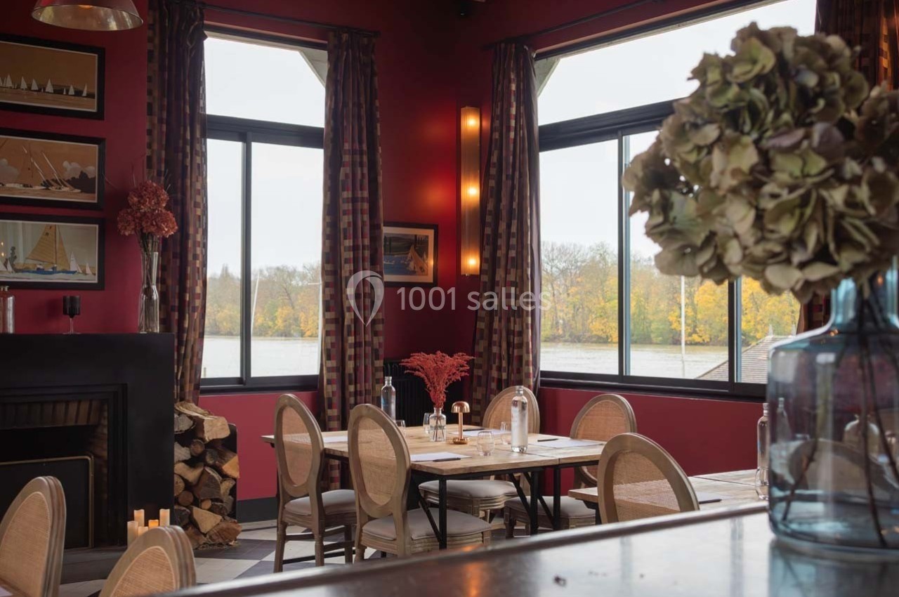 Salle de restaurant chaleureuse avec murs rouges, grandes fenêtres donnant sur un paysage automnal et décoration élégante.