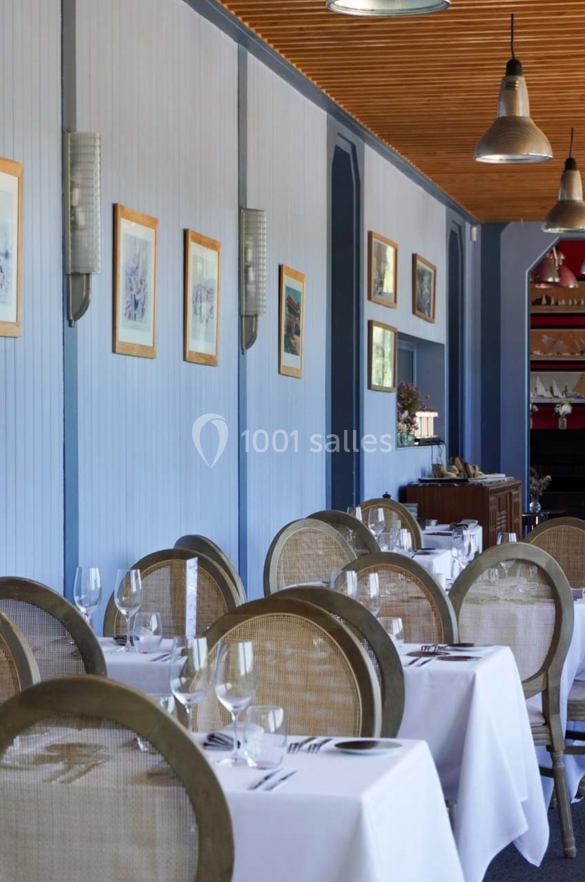 Salle de restaurant élégante avec tables dressées, nappes blanches, chaises en osier et décorations murales.