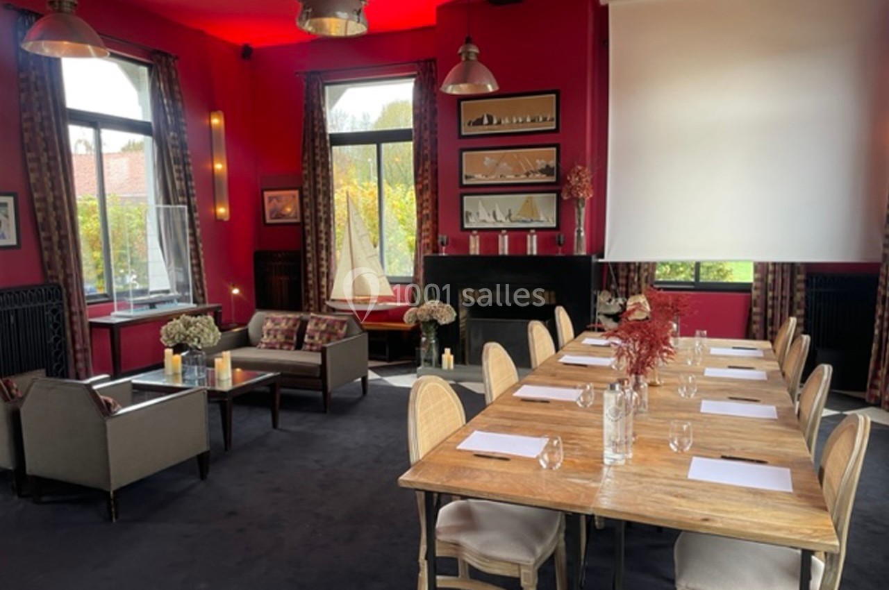 Salle de réunion lumineuse avec murs rouges, table en bois, chaises, coin salon et écran de projection.