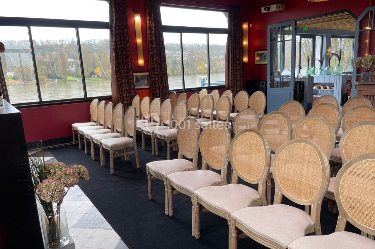Salle aménagée avec des rangées de chaises en bois clair et assises rembourrées, face à de grandes fenêtres donnant sur un…