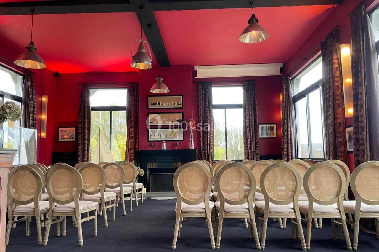Salle de réception lumineuse avec des chaises alignées face à une cheminée, murs rouges et grandes fenêtres.