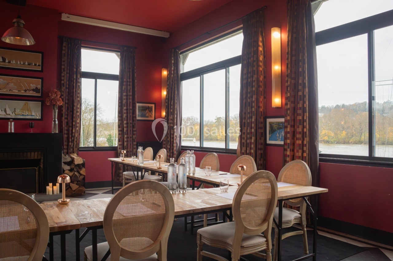 Salle de restaurant lumineuse avec murs rouges, grandes fenêtres, tables en bois et chaises en osier.