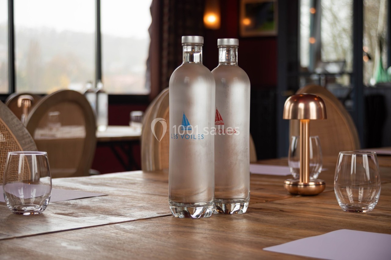 Deux bouteilles d'eau en verre sur une table en bois, entourées de verres et d'une lampe dorée dans un restaurant.