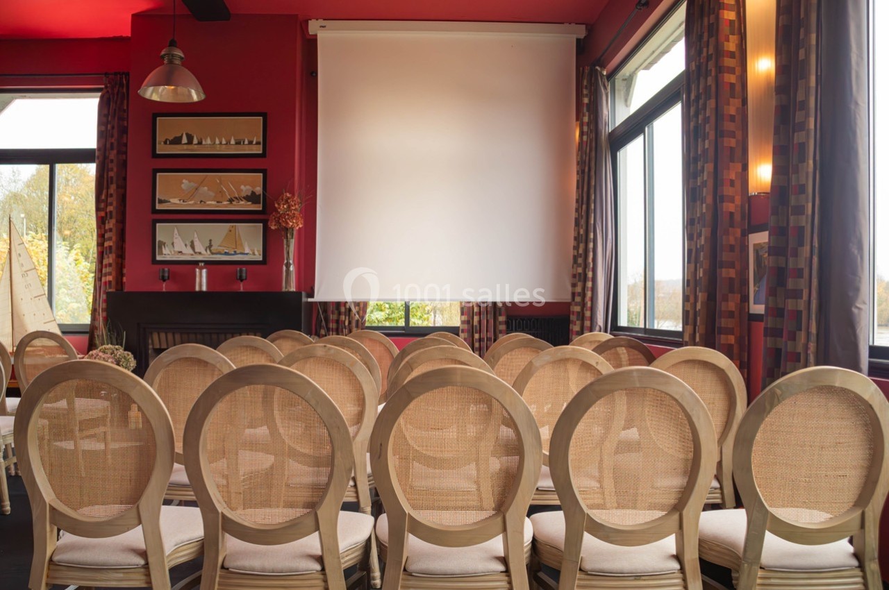 Salle de réunion avec chaises en bois clair alignées face à un écran blanc, entourée de fenêtres et de décorations murales.