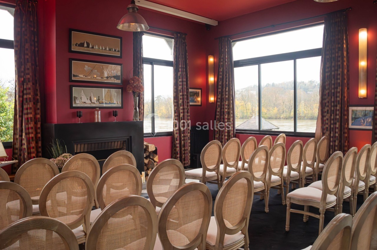 Salle de réunion lumineuse avec chaises alignées, murs rouges, grandes fenêtres donnant sur un paysage arboré.