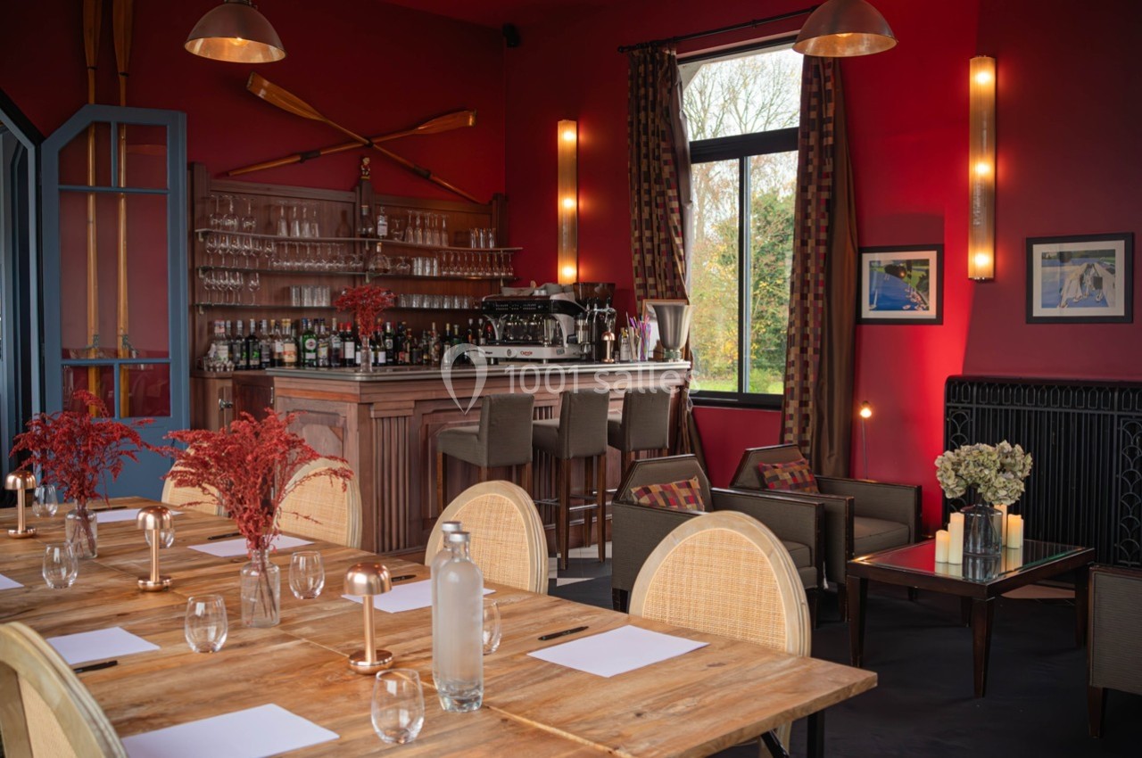 Salle de restaurant chaleureuse avec murs rouges, bar en bois, tables dressées et grandes fenêtres donnant sur l'extérieur.