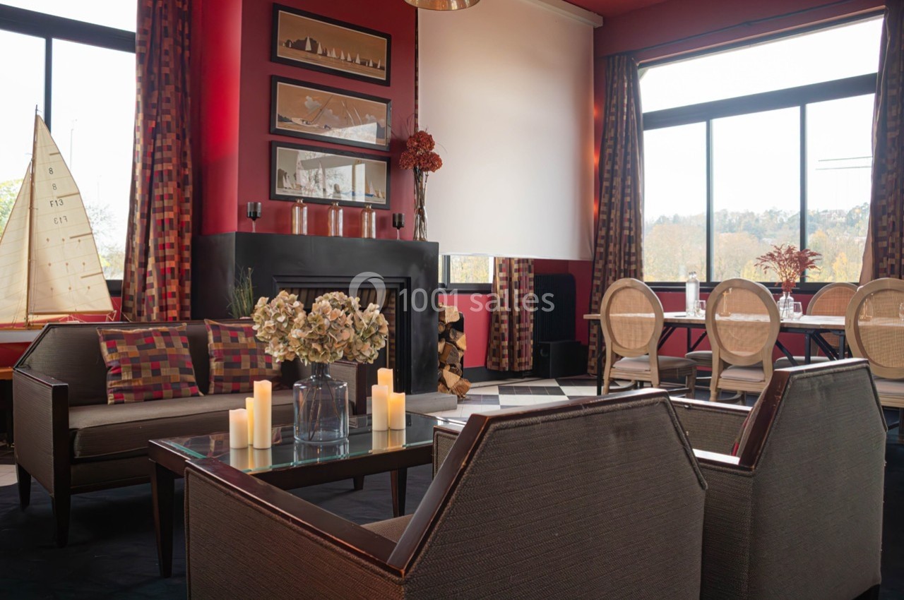 Salon chaleureux avec cheminée, fauteuils, table basse décorée de fleurs et bougies, grandes fenêtres avec vue extérieure.