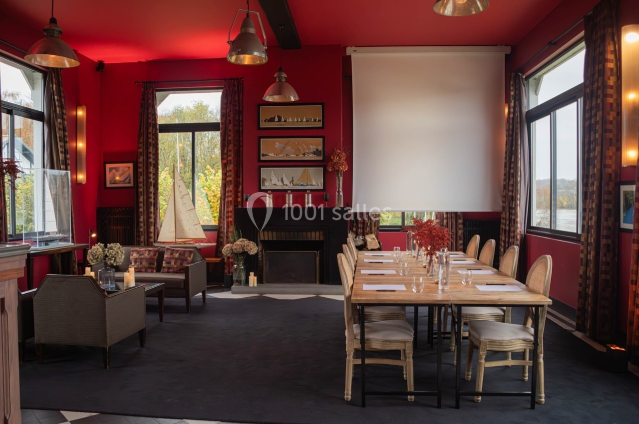 Salle lumineuse avec murs rouges, table dressée pour réunion, cheminée, fauteuils et grandes fenêtres donnant sur l…