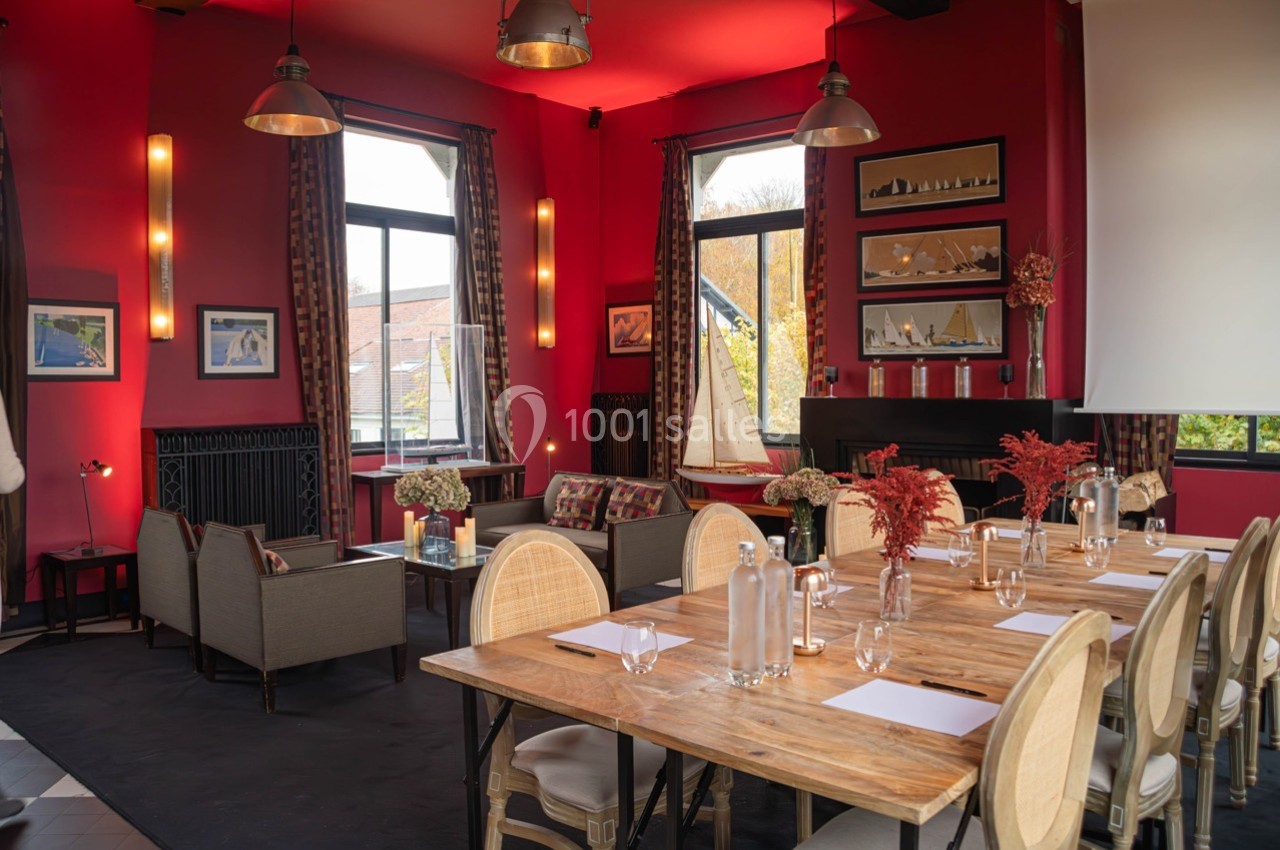 Salle lumineuse avec murs rouges, tables en bois, chaises en rotin, canapés et grandes fenêtres donnant sur l'extérieur.