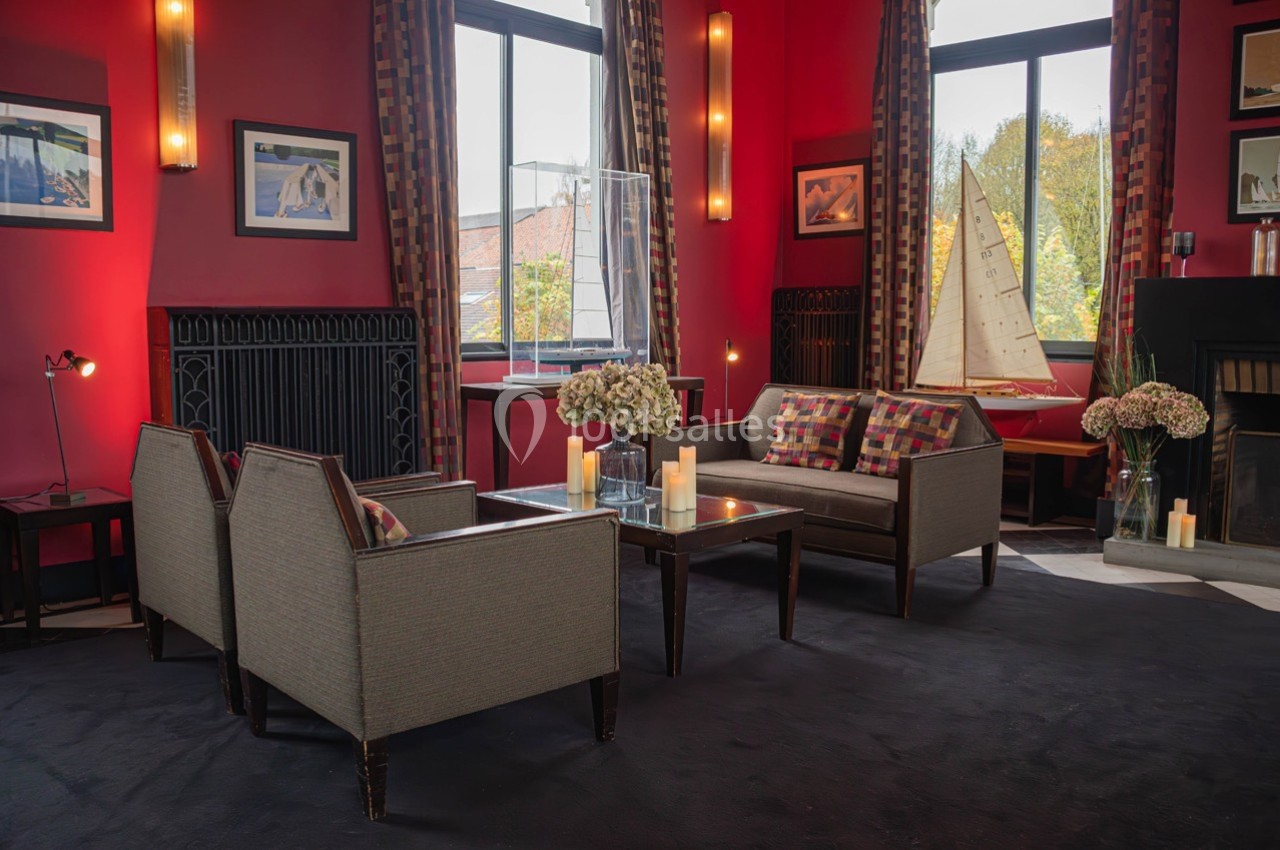 Salon chaleureux avec fauteuils, table basse, bougies, grandes fenêtres, rideaux colorés et décoration nautique.