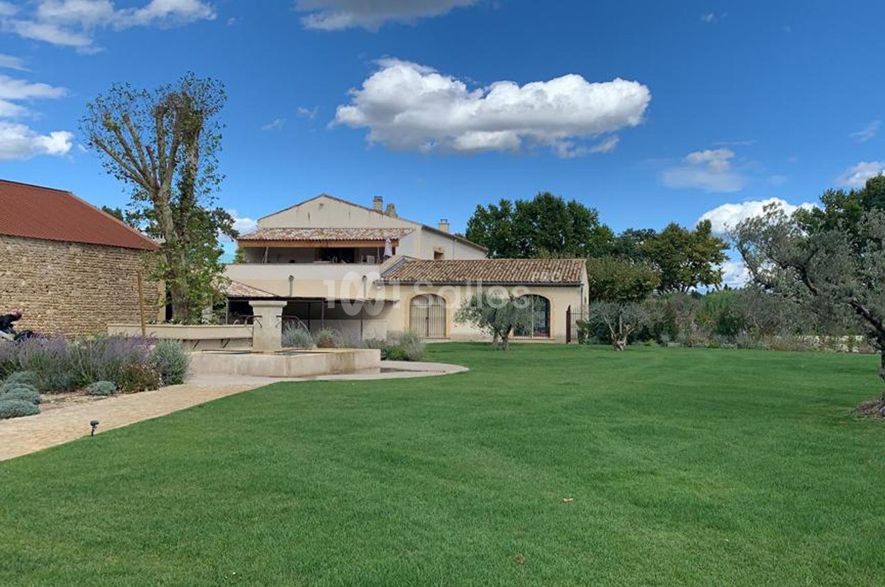 Grande maison provençale entourée de pelouse, d'arbres et d'un ciel bleu avec quelques nuages.
