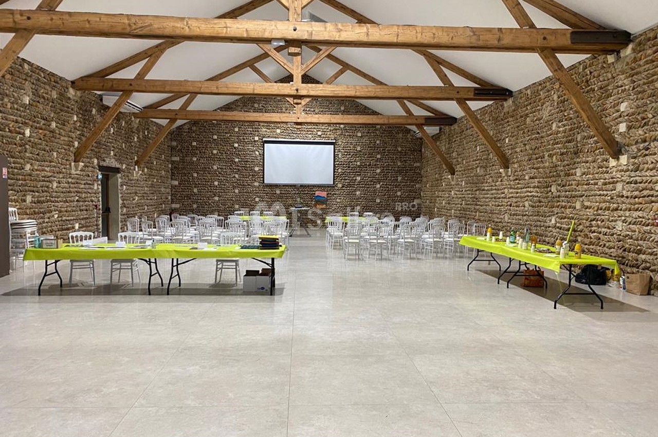 Grande salle en pierre avec poutres apparentes, tables et chaises disposées pour un événement, écran de projection au fond.