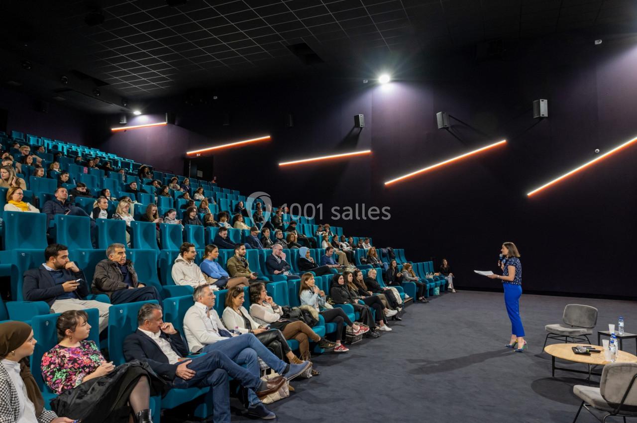 Une conférencière s'adresse à un public assis dans une salle de conférence moderne avec des sièges bleus.