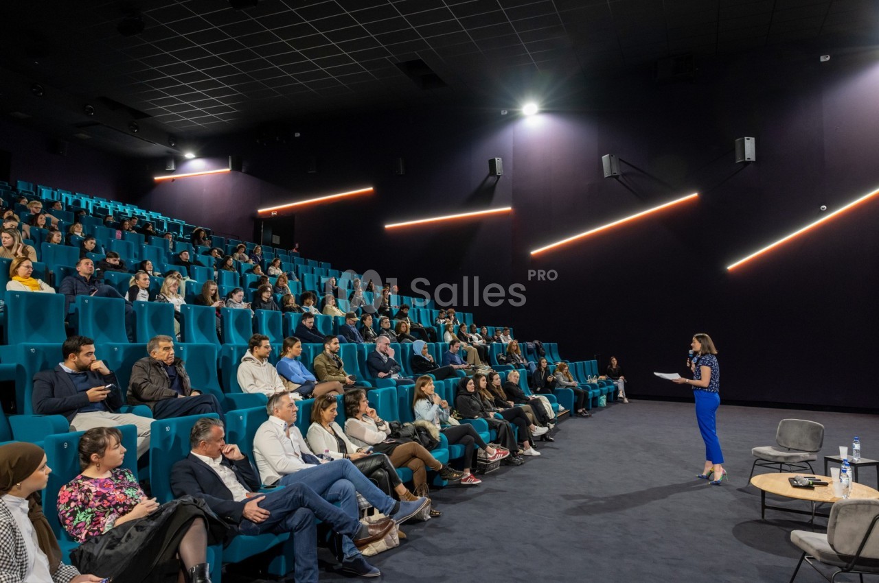 Une conférencière s'adresse à un public assis dans une salle de conférence moderne avec des sièges bleus.