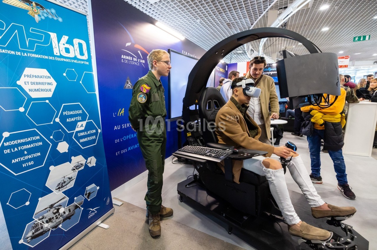 Un homme utilise un simulateur de vol avec un casque de réalité virtuelle, entouré de spectateurs et d'un instructeur en…