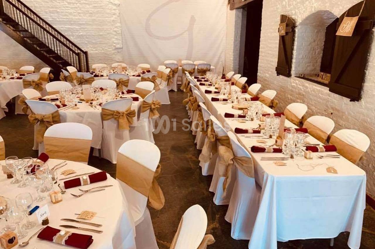 Salle de réception décorée avec des tables dressées, nappes blanches, serviettes rouges et chaises ornées de nœuds beiges.