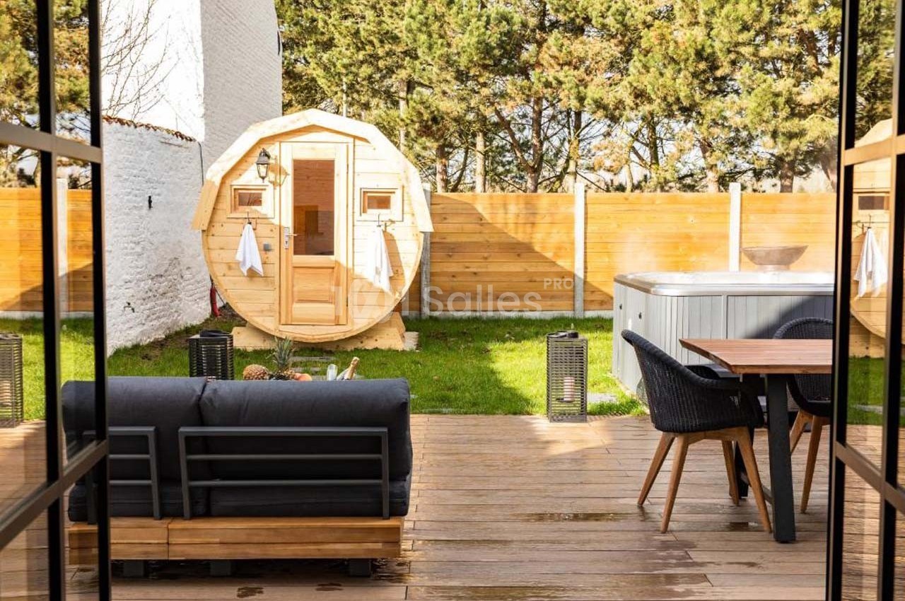 Terrasse en bois avec canapé, table, sauna en forme de tonneau et jacuzzi dans un jardin clôturé.