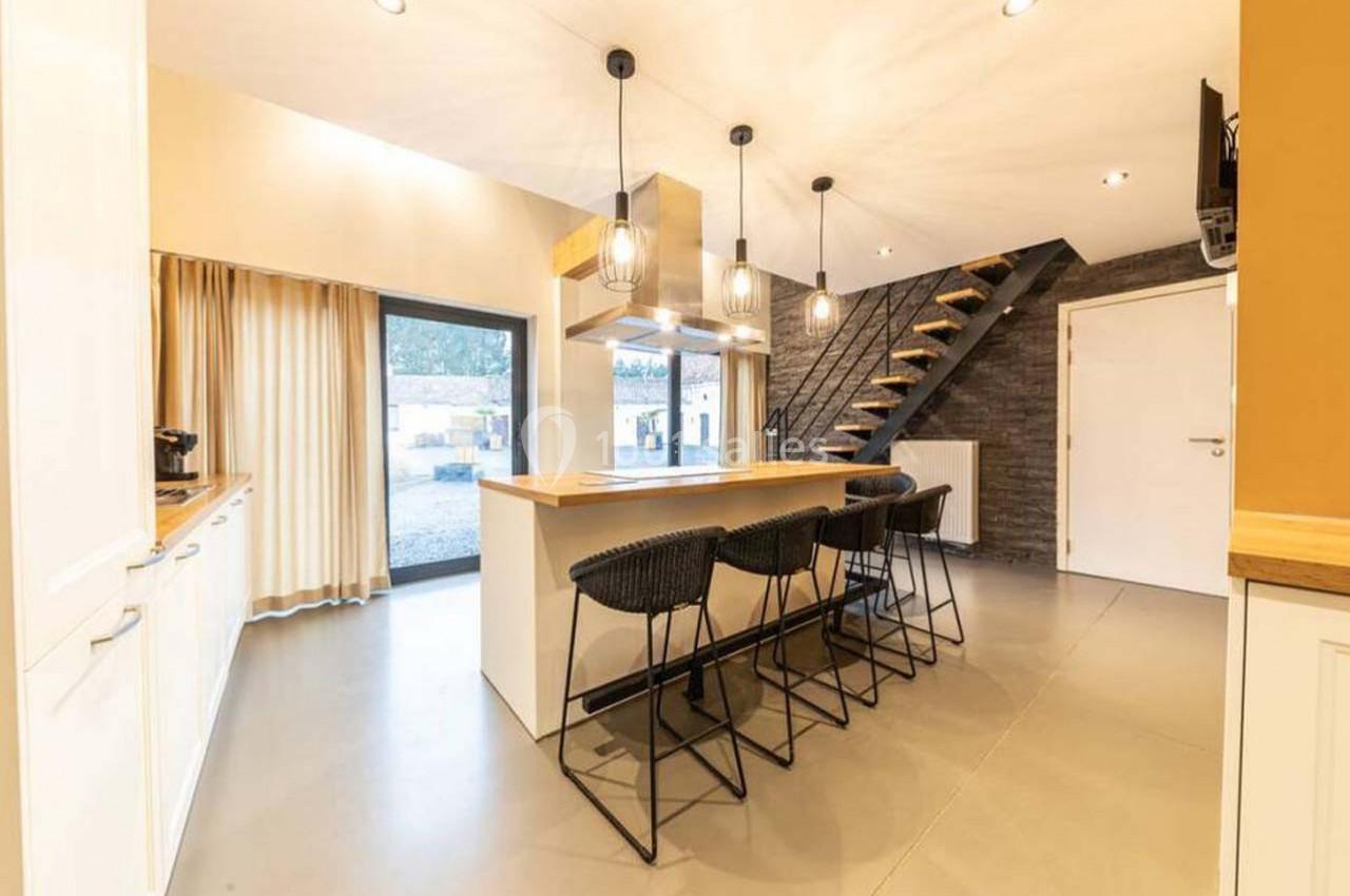 Cuisine moderne avec îlot central, tabourets noirs, éclairage suspendu, escalier en bois et mur en briques sombres.