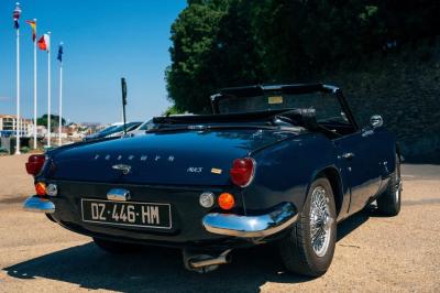 Voiture ancienne décapotable rouge garée sur une pelouse, entourée d'arbres en arrière-plan.