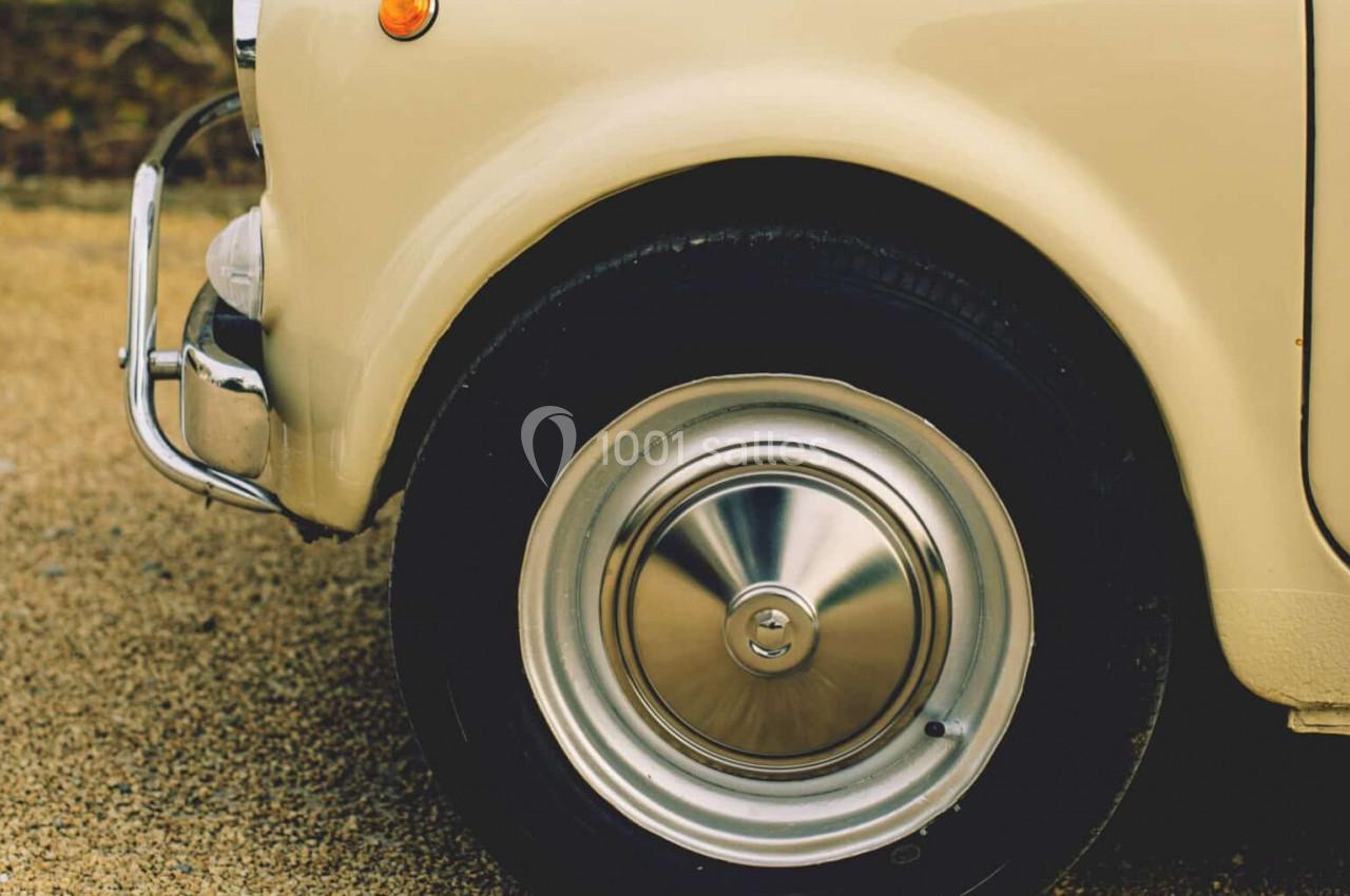 Roue avant d'une voiture vintage beige avec un enjoliveur chromé, stationnée sur un sol gravillonné.