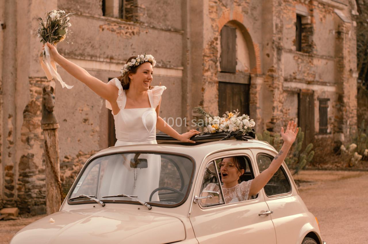 Une mariée souriante agite un bouquet depuis une petite voiture ancienne devant un bâtiment en pierre.