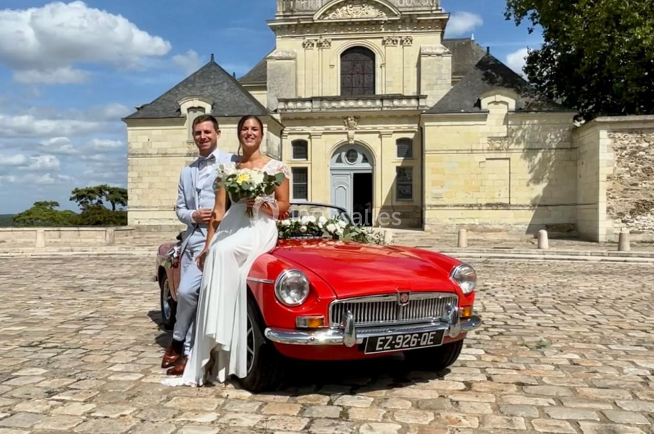 Un couple en tenue de mariage pose devant une voiture rouge ancienne, avec une église en arrière-plan.
