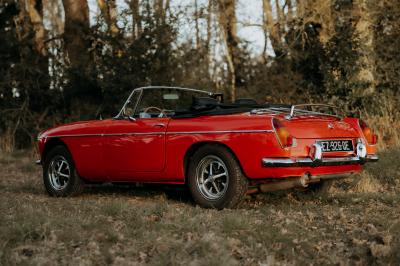Voiture ancienne décapotable rouge garée sur une pelouse, entourée d'arbres en arrière-plan.