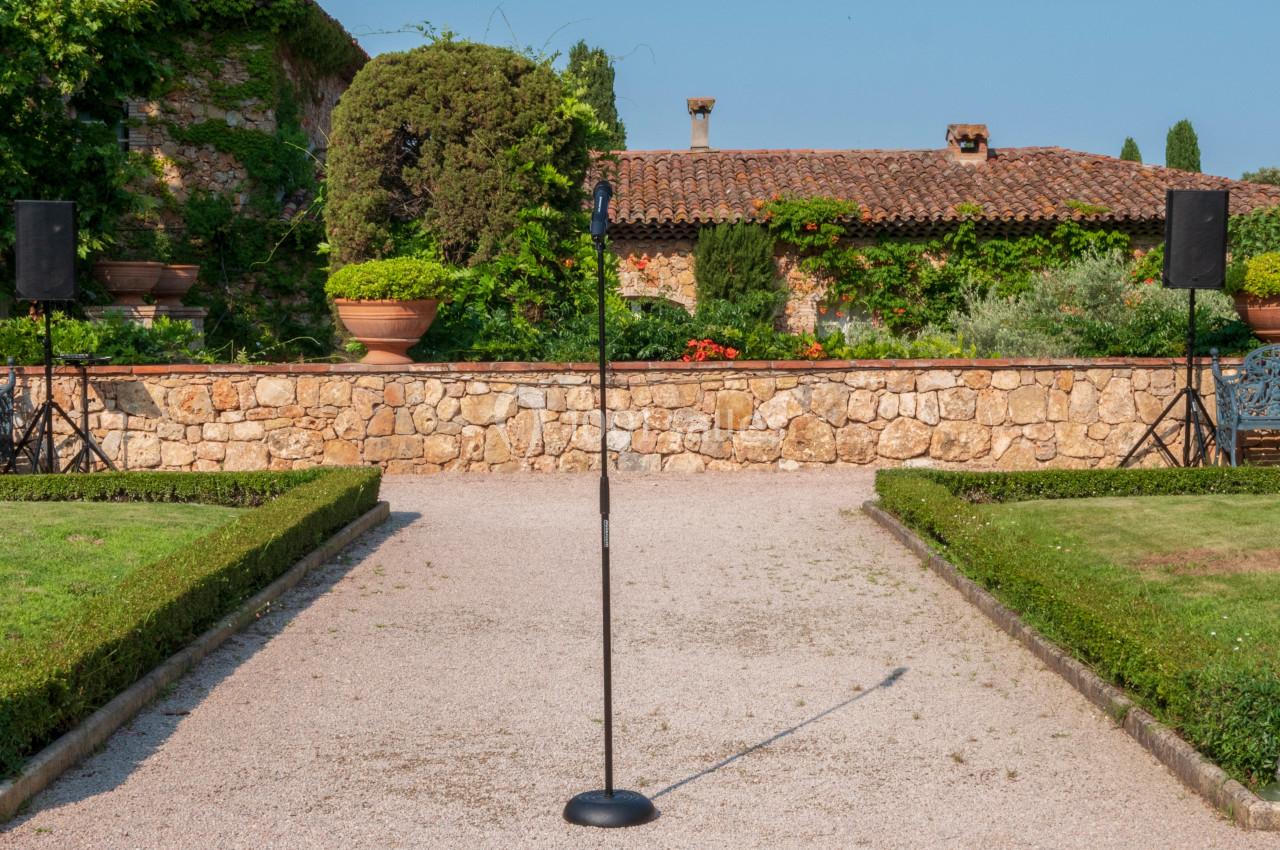 Microphone sur pied installé dans un jardin avec allée centrale, haies taillées et mur en pierre en arrière-plan.