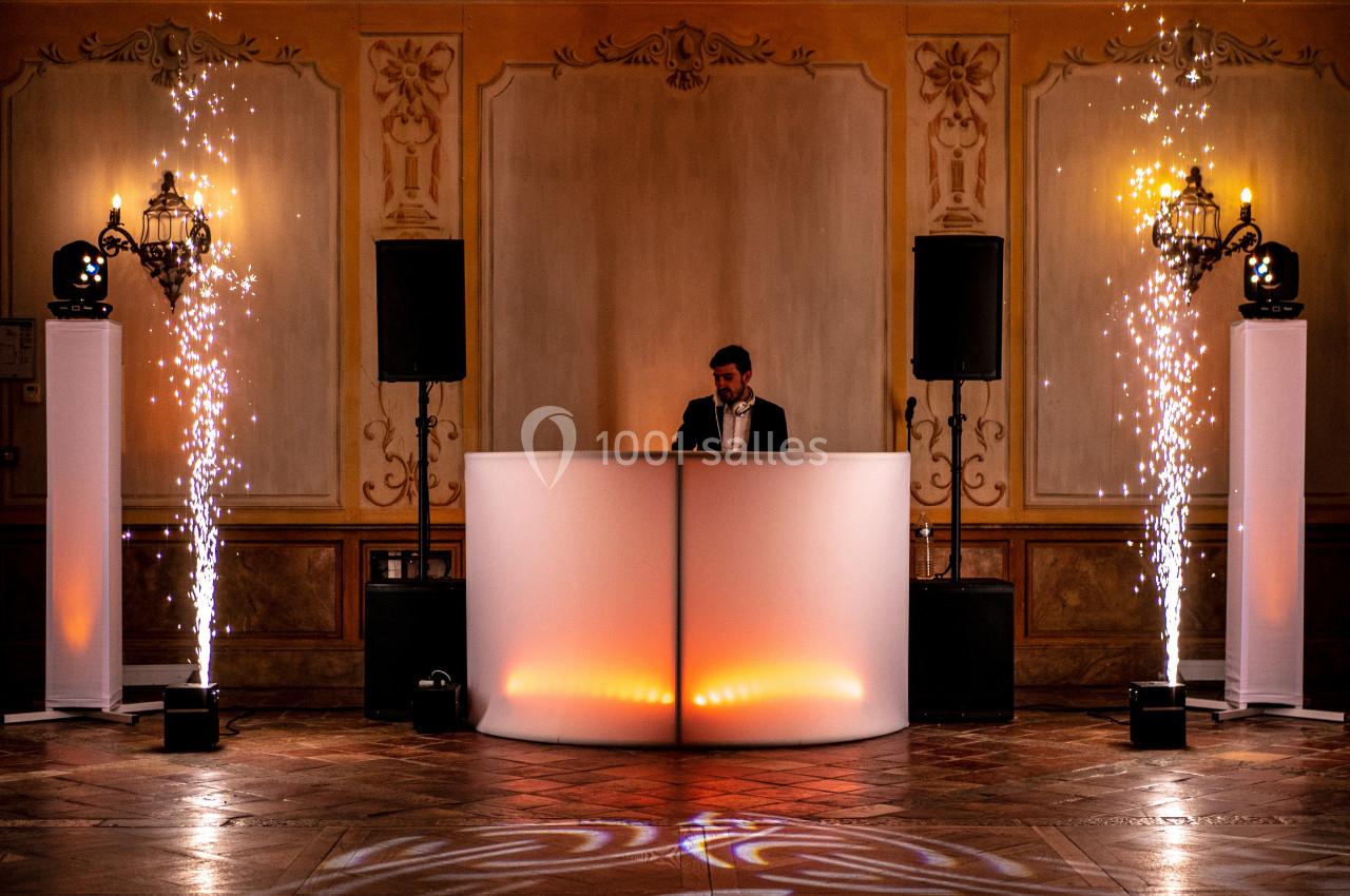 Un DJ mixe derrière une table éclairée, entourée de colonnes lumineuses et d'effets pyrotechniques en intérieur.