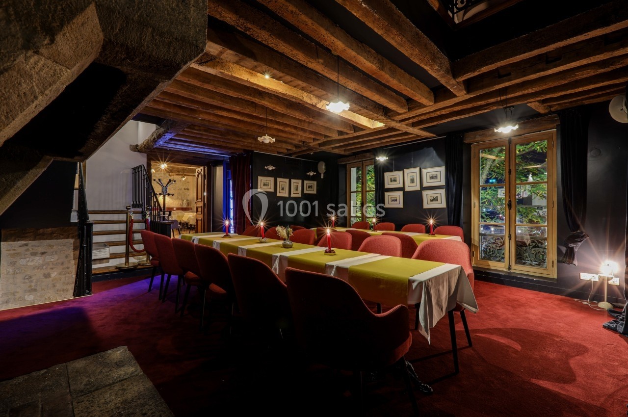 Salle de réunion chaleureuse avec poutres apparentes, tables dressées, chaises rouges et fenêtres donnant sur un jardin.