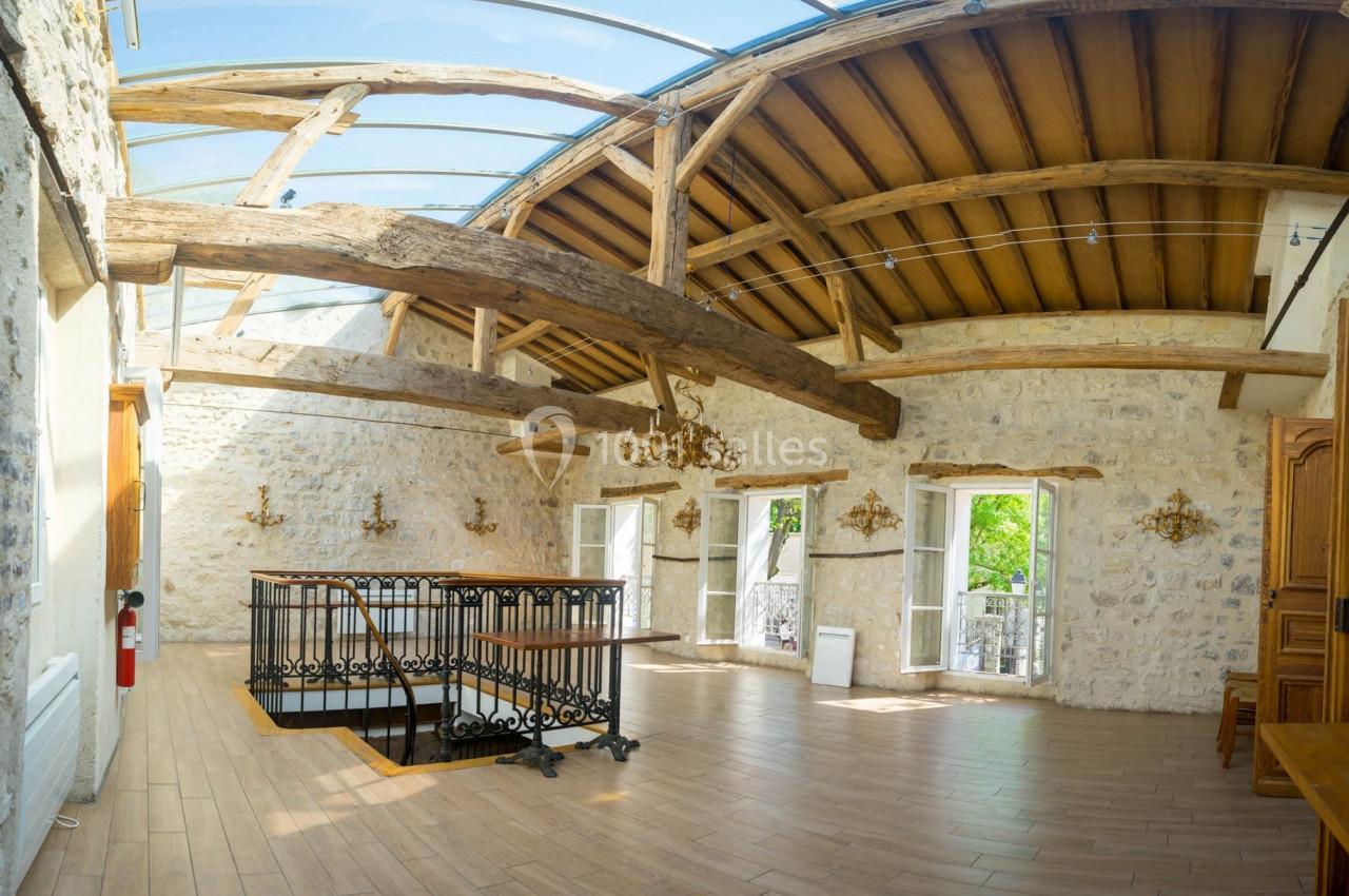 Salle lumineuse avec murs en pierre, poutres apparentes, parquet clair et verrière laissant entrer la lumière naturelle.